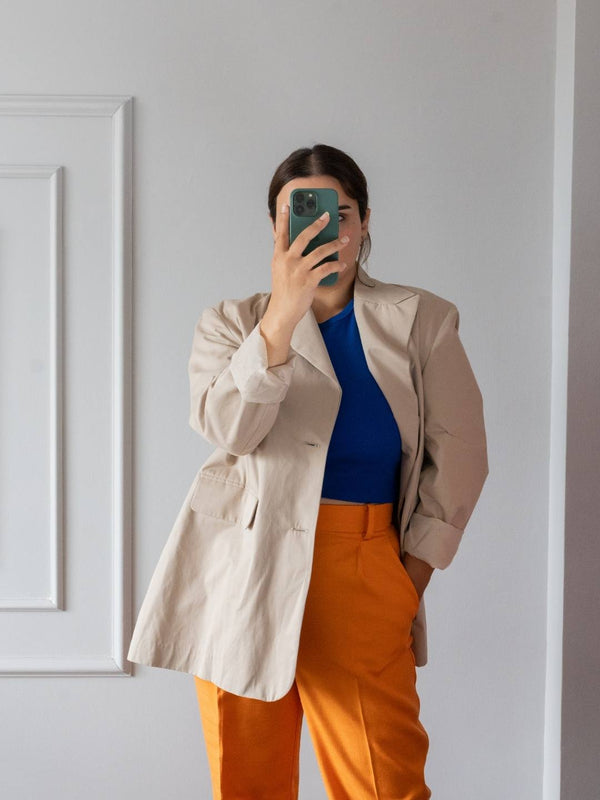 Person wearing a beige blazer, blue top, and orange pants taking a mirror selfie.